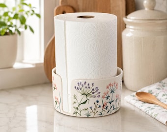 Op de afbeelding: Een cilindrische keukenrolhouder van keramiek met een witte basis versierd met kleurrijke botanische illustraties van wilde bloemen, waaronder paarse, roze en blauwe bloemstengels, met een rol wit keukenpapier met een textuurpatroon.