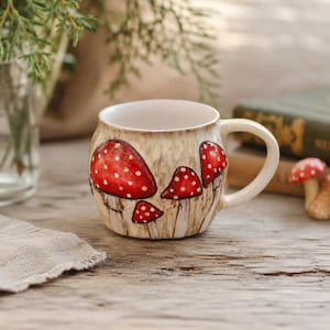 May include: A ceramic mug with a rounded shape and a cream-colored base featuring a hand-painted design of several red mushrooms with white spots. The mug has a single curved handle on the right side and a smooth, glossy finish throughout.