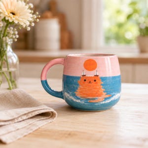 Peut inclure: Une tasse en céramique peinte à la main représentant un chat orange nageant dans une eau bleue avec des vagues blanches. L'arrière-plan montre un ciel rose avec un soleil orange vif et deux petits voiliers. La tasse a une anse rose et bleue.