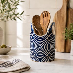 Può includere: Un portautensili cilindrico in ceramica con base bianca e un motivo ripetitivo di spirali e cerchi blu scuro. Il contenitore è dotato di due manici laterali integrati e contiene due cucchiai in legno posizionati in verticale all'interno.