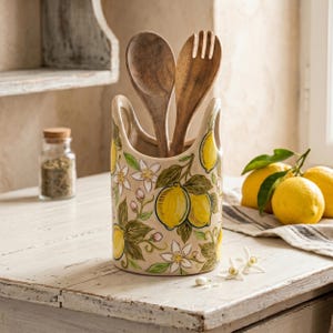 May include: A cylindrical ceramic utensil holder with a cream-colored base featuring a hand-painted pattern of yellow lemons, green leaves, and white blossoms. Two wooden kitchen utensils, a spoon and a fork, are placed inside the container.