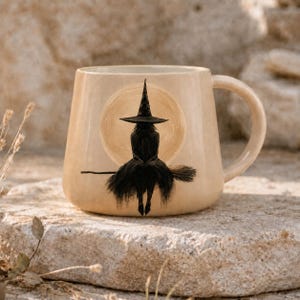May include: A beige ceramic mug featuring a black silhouette illustration of a figure wearing a pointed hat and sitting on a broomstick, centered against a pale yellow circular moon design. The mug has a smooth finish and a curved handle.