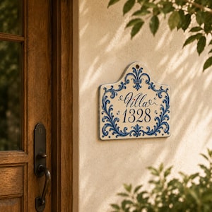 May include: A cream-colored ceramic house sign with a decorative blue scrollwork border. The sign features the text "Villa" in elegant script above the number "1328" in a classic serif font, with a small red heart accent between the words.