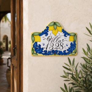 May include: A rectangular ceramic house sign with a curved top, featuring a hand-painted design of yellow lemons and green leaves. The center displays the black text Villa 1328, surrounded by intricate blue scrollwork on a white background.
