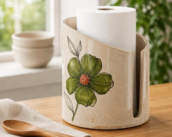 Op de afbeelding: Een cilindrische keukenrolhouder van keramiek met een gespikkelde beige afwerking en een handgeschilderd groen bloemmotief met een textuurrijk oranje hart. De houder heeft een verticale uitsparing aan de rechterkant voor toegang tot de rol.