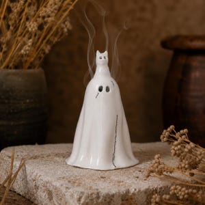 Könnte beinhalten: Ein weißer Keramik-Räucherstäbchenhalter in Form eines Geistes mit Katzenohren. Die Figur hat zwei kreisförmige Augenausschnitte und eine vertikale, schwarz aufgemalte Nahtlinie an der Seite. Rauch steigt aus dem Kopf und den Augenöffnungen auf.