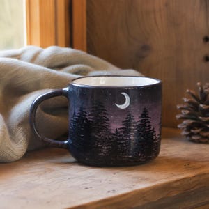 May include: A ceramic mug featuring a hand-painted night scene with a dark purple and black gradient sky, a white crescent moon, and a silhouette of pine trees. The mug has a smooth, rounded handle and a white interior finish.