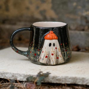 May include: A black ceramic mug featuring a raised, three-dimensional white ghost figure wearing an orange beanie. The mug is decorated with a painted field of small red and yellow flowers that wrap around the base, set against a dark background.