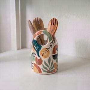 Pode incluir: Um porta-utensílios cilíndrico de cerâmica com um padrão abstrato pintado à mão em tons de azul-petróleo, terracota, amarelo-mostarda e preto. O design inclui formas orgânicas, motivos de folhas e um ícone de olho. Dois talheres de madeira estão dentro.