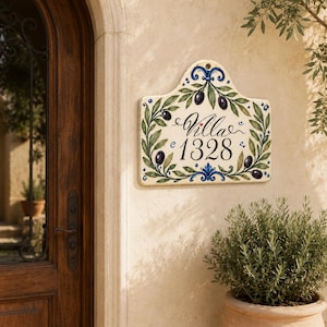 May include: A rectangular ceramic wall plaque with a curved top edge, featuring a hand-painted design of green olive branches with dark purple olives. The center displays the black text Villa 1328 with a small red heart above the letter i.
