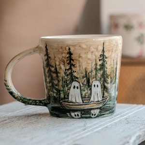 Peut inclure: Une tasse en céramique avec un fond beige présentant une scène peinte à la main de deux fantômes blancs assis dans un petit bateau sur un lac sombre. Le paysage environnant comprend de grands pins verts et le reflet des fantômes dans l'eau.