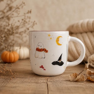 Könnte beinhalten: Ein weißer Keramikbecher mit handgemalten Illustrationen eines Geistes mit orangefarbenem Schottenkaro-Schal, einem gelben Halbmond, einem schwarzen Hexenhut, kleinen roten Pilzen und goldenen Sternen auf der Außenfläche.
