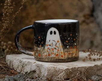 Op de afbeelding: Een keramische mok met een glanzend zwarte buitenkant, voorzien van een handgeschilderde witte spookfiguur die in een veld met kleine oranje en gele bloemen staat. De mok heeft een gebogen handvat en een witte binnenkant met een artistiek ontwerp.
