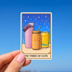 May include: A sticker featuring an illustration of a pink water bottle, a glass of iced coffee with a straw, and a yellow can. The background is a starry night sky. The text "THE THREE OF CUPS" is at the bottom.