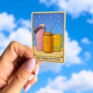 May include: A tarot card illustration featuring a pink water bottle, a jar of iced tea with a straw, and a yellow can. The card is titled "THE THREE OF CUPS" against a starry blue background.