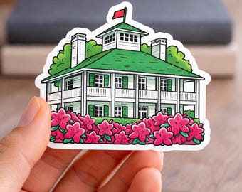 May include: A sticker featuring a white house with a green roof and shutters, a red flag, and pink flowers. The house has multiple windows and a porch. The sticker is held in a hand.