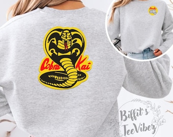 May include: Grey sweatshirt with a yellow Cobra Kai logo featuring a black cobra with red eyes and the words "Cobra Kai" in red. The sweatshirt is shown on a model.