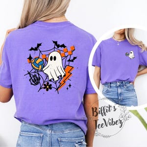 May include: A purple t-shirt featuring a Halloween-themed graphic. The design includes a white ghost, a volleyball, bats, a spiderweb, and a lightning bolt. The shirt has a crew neck and short sleeves.