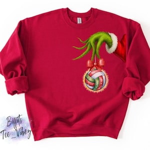 May include: Red sweatshirt featuring a green hand with a red and white trimmed sleeve holding a volleyball ornament. The volleyball ornament has a red bow and is decorated with green, white, and red stripes. The sweatshirt has long sleeves.