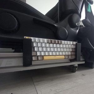 May include: A white mechanical keyboard with gold keycaps is mounted on a black racing seat. The keyboard is attached to the seat with a black bracket. The seat is on a gray floor.
