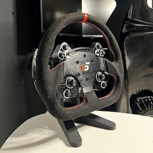 Può includere: Un volante da corsa nero con cuciture rosse e un supporto nero. Il volante ha un design in fibra di carbonio e un logo #5 rosso e arancione.