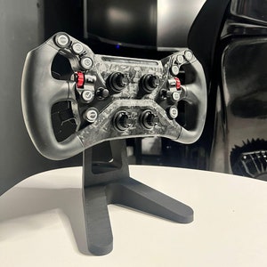 May include: A black racing steering wheel with a carbon fibre design and multiple buttons and switches. The steering wheel is mounted on a black stand.