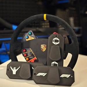 May include: A black racing steering wheel with yellow accents and various buttons and controls. The wheel is attached to a black and silver base with a Porsche logo. There are three black paddle shifters with white logos: a racing driver, "F1", and a logo with a stylized fist.