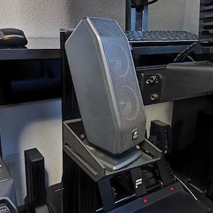 May include: A black computer speaker mounted on a black metal stand with a carbon fiber pattern. The speaker has a black grill and a black and silver logo. The stand has three red lights on the front.