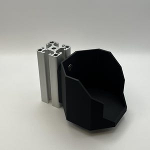 May include: A black, multi-sided, open-top container attached to a silver, square-profile aluminum extrusion rail. The container features a flat base and vertical walls, with a single visible metal screw securing it to the metal rail structure.