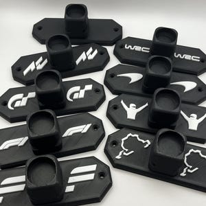 May include: A collection of black wall mounts featuring various racing logos, such as WEC, GT, F1, and more. Each mount has a circular cutout designed to hold a racing trophy or other collectible.