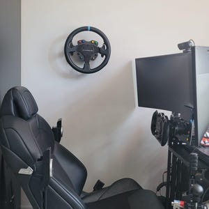 May include: A black racing simulator setup featuring a mounted steering wheel with colored buttons and a black racing seat. A large monitor and additional controls are also visible, creating an immersive gaming environment.