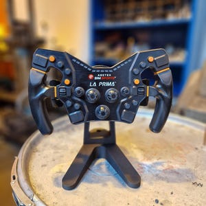 May include: Black and orange racing simulator steering wheel with the text "LA PRIMA" and "ASETEK SIMSPORTS". The wheel has multiple buttons and dials and is mounted on a black stand.