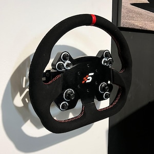 May include: A black racing steering wheel with red stitching and a red stripe on the top. The wheel has a black and white checkered flag logo with the number "5" in the center. The wheel is mounted on a white wall.