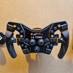 May include: A black and orange racing simulator steering wheel with the text "ASETEK SIMSPORTS LA PRIMA". The wheel has multiple buttons and dials, with orange accents on the handles and buttons. The wheel is mounted on a light brown surface.