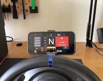 May include: A black racing steering wheel with a blue stripe and a smartphone mounted above it. The phone screen displays a racing game interface with data like lap time, oil temperature, and tire pressure. The screen also shows an alarm for low fuel pressure.
