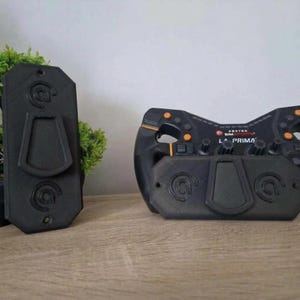 May include: A black Asetek SimSports La Prima steering wheel with orange buttons and a matching black mounting plate. The steering wheel features a central display area with the text Asetek SimSports La Prima and multiple rotary knobs.
