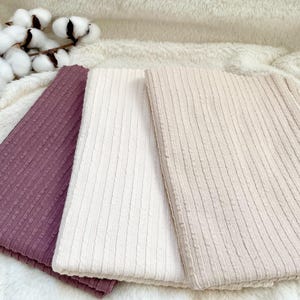 Può includere: Tre pezzi di tessuto a coste piegati, nei toni del malva, crema e beige, sono esposti su una soffice superficie bianca. Rami di cotone sono disposti nell'angolo in alto a sinistra. Il tessuto ha un motivo a coste verticale strutturato.