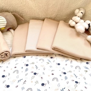 Puede incluir: Un conjunto de artículos para bebés, que incluye mantas plegadas en varios tonos y texturas beige, una manta enrollada y un juguete de elefante de peluche. Se ve una sábana estampada con caracoles, arcoíris y hojas.