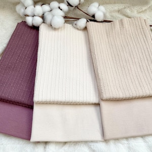 Può includere: Tre pezzi di tessuto piegati nei toni malva, crema e beige. Gli strati superiori hanno una trama a coste, gli strati inferiori sono lisci. Rami di cotone con fiori bianchi sono sullo sfondo. I tessuti sono probabilmente per cucire o creare.