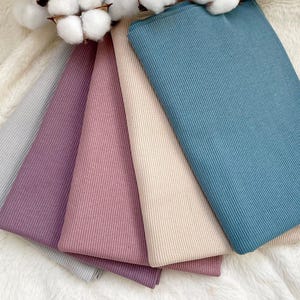 Può includere: Una collezione di tessuti a maglia a coste colorati in tonalità di grigio, viola, rosa, beige e verde acqua. I tessuti sono disposti a ventaglio, mostrando la loro consistenza e le variazioni di colore. Rami di cotone sullo sfondo.