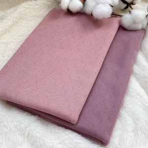 Può includere: Due pezzi di tessuto piegati, rosa e viola, con un piccolo motivo a rombi, sono presentati su una superficie bianca e soffice. Rami di cotone sono posti in alto, aggiungendo un elemento naturale alla presentazione.