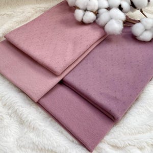 Può includere: Quattro quadrati di tessuto piegati in tonalità rosa e viola sono disposti su una superficie bianca e soffice. Gli strati superiori presentano un delicato motivo a pois. Un rametto di cotone aggiunge un accento naturale.