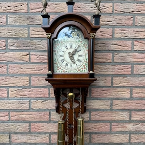 May include: Antique wooden wall clock with a Roman numeral face, ornate carvings, and three brass figures on top. The clock has a pendulum and two brass weights hanging below.