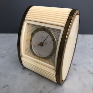 May include: A white and gold vintage alarm clock with a square face and a grid pattern. The clock face has black hands and the word "VEGLIA" printed on it.