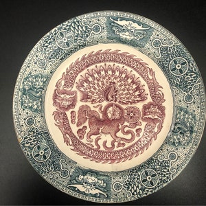 May include: Decorative ceramic plate with a detailed Asian-inspired design. The plate features a central maroon image of a mythical creature surrounded by a dragon and peacock motif. The border is teal with repeating patterns.