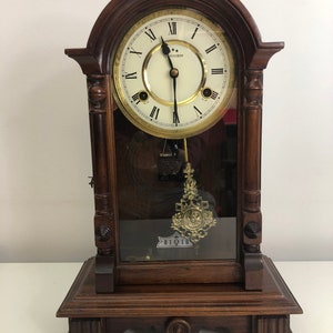 Puede incluir: Un reloj antiguo de madera oscura con una cara de vidrio y un péndulo. La cara del reloj tiene números romanos y las palabras "Commodor" y "Big Ben".
