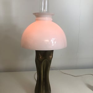 May include: A table lamp made from a repurposed brass shell casing. The lamp has a white glass shade with a pink glow and a clear glass chimney.