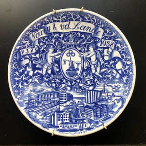 Puede incluir: Un plato de cerámica azul y blanco con un diseño decorativo que representa una escena de fábrica, dos ángeles y el texto "Noury & vd. Lande 1838 1938".