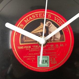Op de afbeelding: Een zwarte vinyl platenspeler klok met witte wijzers. De wijzerplaat heeft een rood label met gouden letters met de tekst "His Master's Voice" en "Tone-Poem "Finlandia" Op. 26, No. 7 (2nd Record) Leopold Stokowski The Philadelphia Orchestra (42-1174) Speed 78 Cat. No. D.B. 1584" Het label heeft ook een zwart-witte illustratie van een grammofoon en een hond.