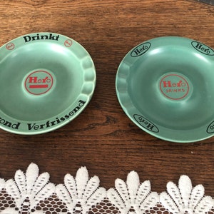 Könnte beinhalten: Zwei grüne Aschenbecher aus Keramik mit dem Wort "Hero" und dem Text "Drinkt Gezond Verfrissend" auf einem Aschenbecher und "Hero Drinks" auf dem anderen.